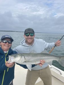 Capt. Avery holding a striped bass in front of a client holding a fly rod